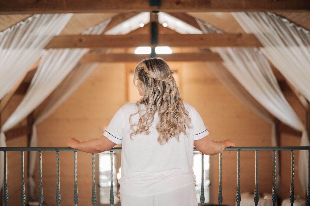 Bride is looking to the right, her hair in a half up half down look. There is ceiling drapery behind her in a barn venue.